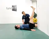 Ein Trainer hilft einem Mann beim Oberschenkel dehnen. Der Mann liegt auf dem Rücken, das Bein gestreckt an der Wand.