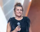 Uśmiechnięta finalistka, która mogła wygrać Voice Senior, w czarnej, błyszczącej sukni z futrzanym kołnierzem, trzyma mikrofon.