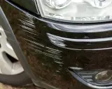 Pęknięty lakier na zderzaku czarnego samochodu, pełen licznych rys. Widać fragment reflektora i felgi.