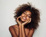 Una mujer sonríe con los ojos cerrados, su cabello rizado enmarca su rostro. Su piel radiante y su expresión serena sugieren cómo parecer más joven.