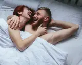 Para kochająca się w łóżku, śmiejąca się. Kobieta z rudymi włosami obejmuje mężczyznę z brodą. Sikanie podczas seksu może być częścią intymnych chwil.