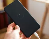 Dłoń trzyma smartfon z ikoną ładowania zwrotnego na czarnym ekranie. Podłączony biały kabel USB.