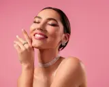Mujer sonríe con ojos cerrados, luciendo un maquillaje romántico, joyas brillantes y un fondo rosa.