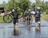 Kolarze pokonują wodną przeszkodę podczas mazowiecki gravel. Niosą rowery przez płytką rzekę, w tle zielone drzewa i samochód.