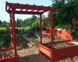 A red wooden pergola frame stands in a garden with raised beds and greenery.