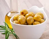 Aceitunas verdes en un cuenco blanco junto a una botella de aceite de oliva y romero. Descubre los aceitunas beneficios y contraindicaciones.