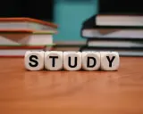 Białe kostki z literami "STUDY" na drewnianym stole, w tle stosy książek.