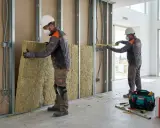 Dos obreros instalan aislamiento en una pared. El material, que parece lana de roca, se coloca entre perfiles metálicos. Se ve una caja de Rockwool y herramientas. Las subvenciones se devuelven con trabajo de calidad.