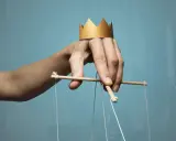 Dłoń z papierową koroną na palcach, sterująca marionetką. Symbol władzy i despoty.