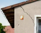 Grieta en pared de edificio, indicando problemas de **seguridad estructural**. Se observa un alero de madera y un cielo azul.