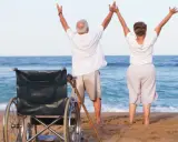 Szczęśliwi seniorzy cieszą się urlopem. Wózek inwalidzki i laska na plaży świadczą o tym, że to udany turnus rehabilitacyjny nad morzem.
