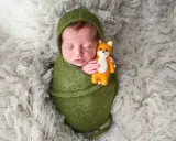 Dolci immagini neonati: un bimbo avvolto in una coperta verde tiene stretto un piccolo volpino di peluche, dormendo sereno su un soffice tappeto bianco.