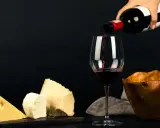 Vin rouge versé dans un verre, à côté de morceaux de fromage. La question "quoi boire avec une raclette" trouve ici une réponse évidente.