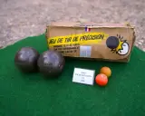 Deux boules de pétanque et un cochonnet sur un tapis vert. La boîte du jeu de tir de précision est visible. Apprenez comment tirer à la pétanque !