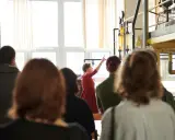 Grupa studentów na zajęciach praktycznych. Kobieta w czerwonej sukience tłumaczy coś przy aparaturze. To może być część ich studiów magisterskich zaocznych w Warszawie.