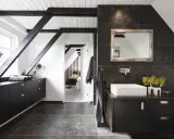 Baño moderno con vigas oscuras y suelo gris. El lavabo sobre encimera y los muebles oscuros contrastan con las paredes de baldosas oscuras. Se ve un dormitorio al fondo.