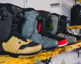 Rząd butów snowboardowych w różnych kolorach i stylach, ustawionych na drewnianej półce.