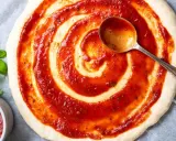 Pizzaboden mit selbstgemachter Pizzasauce bestreichen. Frisches Basilikum und ein Löffel mit Sauce daneben.