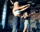 Ein Paar trainiert mit Kettlebells im Fitnessstudio. Sie heben Gewichte, während eine dritte Person im Hintergrund sitzt.