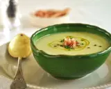 Un refrescante gazpacho de melón, adornado con jamón serrano y cebollino, servido en un cuenco verde con una cuchara dorada.