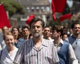Nanni Moretti cammina in una folla con bandiere rosse, sullo sfondo il Colosseo.