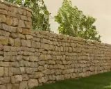 Un muret en pierre sèche, bordé d'herbe verte et de verdure luxuriante, s'étend sur la droite, menant à des marches en pierre.