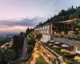 Un lussuoso hotel si affaccia sulle **colline di Firenze** al tramonto, con giardini illuminati e la città in lontananza.