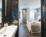 Badezimmer mit Waschbecken und Blick auf ein Hotelzimmer mit Bett und Fenstern.