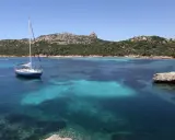 Un voilier bleu ancré dans les eaux turquoise de Cap Corse, avec des collines verdoyantes en arrière-plan sous un ciel clair.