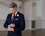 Elegancki mężczyzna w granatowym garniturze i białej koszuli, z bordowym krawatem i butonierką, przygotowuje się na wesele.