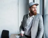 Mężczyzna w szarej czapce snapback i garniturze trzyma piłkę baseballową.