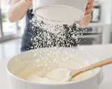 Une main saupoudre du glacage sucre glace sur une préparation crémeuse dans un bol blanc, avec une cuillère en bois.