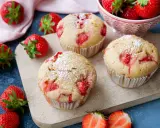 Saftige Erdbeer Muffins, bestäubt mit Puderzucker, liegen auf einem Holzbrett, umgeben von frischen Erdbeeren.