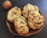 Quatre pains aux lardons et oignons dorés dans un panier en osier, avec un oignon cru à côté.
