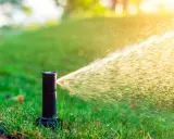 Aspersor de riego automático en acción, rociando agua sobre césped verde bajo la luz del sol.