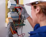 Elektryk w białym kasku przeprowadza przegląd instalacji elektrycznej, mierząc napięcie multimetrem.
