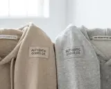 Dwa bluzy z kapturem, beżowa i szara, z naszywką "AUTHENTIC GOODS CO." i metką "100% ORGANIC COTTON | MADE IN JAPAN". Oryginalna metka Champion.
