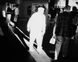 A stark black and white image by Trent Parke captures a blurred figure in a white hat and coat, walking with a cane. Other figures are also blurred, suggesting movement and a fleeting moment.