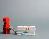 Czerwony ludzik w kajdankach, symbolizujący więzienie za długi, stoi obok stosów pieniędzy.