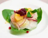 Carpaccio de saint jacques frais, garni d'orange, d'oignon rouge et de pousses vertes, sur une feuille d'épinard.