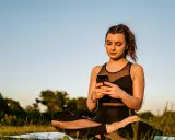 Kvinna i lotusställning på en matta utomhus, med en mobiltelefon i händerna. En modern typ av meditation.