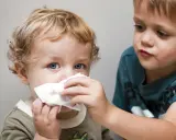 Starszy brat pomaga młodszemu z katarem u niemowlaka. Chłopiec podaje chusteczkę, by otrzeć nosek maluchowi.