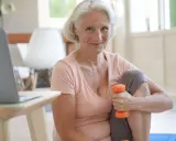 Frau über 60 mit Hanteln zu Hause für Muskelaufbau. Sie lächelt und trainiert vor ihrem Laptop.