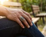 Un uomo indossa un anello con croce, simbolo di fede e protezione. Il sole illumina la sua mano poggiata sui jeans.