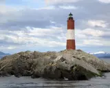 Faro a strisce rosse e bianche su un'isola rocciosa nel Canale di Beagle, con montagne innevate sullo sfondo.