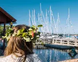 En flicka med blomsterkrans ser ut över en hamn med segelbåtar. En perfekt dag för en resa till Åland.