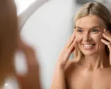 Donna sorride applicando crema contorno occhi. La sua pelle sottile del viso mostra segni del tempo, ma è luminosa.