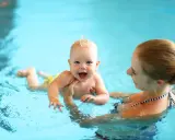 Glückliches 8 Wochen altes Baby mit Mutter im Schwimmbad. Das Baby lacht und planscht im Wasser, während die Mutter es festhält.
