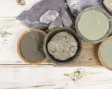 Pots de différentes argiles, dont une argile verte pour dégonfler, sur un fond en bois blanc et brut.
