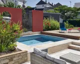 Une piscine moderne avec une belle décoration autour piscine : mur rouge, terrasse en bois, mobilier de jardin et végétation luxuriante.